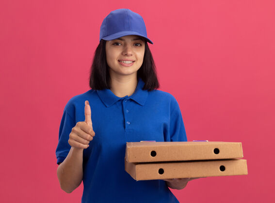 站着身穿蓝色制服的年轻送货女孩手持披萨盒 友好地站在粉红色的墙上 竖起大拇指微笑着盒子抱着微笑