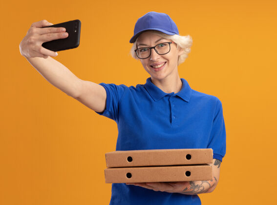 用着身穿蓝色制服 戴着眼镜 拿着比萨饼盒 用智能手机在橙色墙上微笑的年轻送货员拿着帽子制服