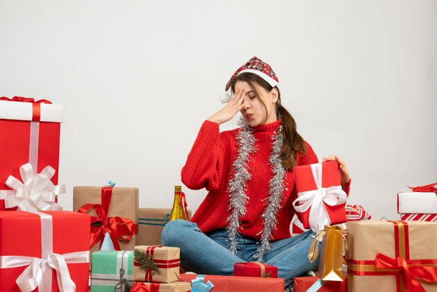 坐着前视图困惑的女孩拿着圣诞帽拿着礼物坐在礼物周围周围礼物圣诞老人