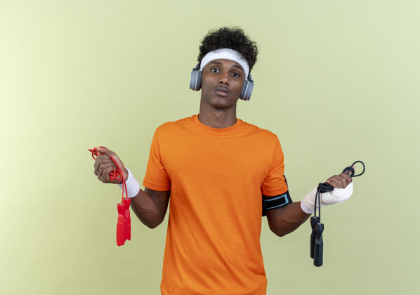 举行困惑的年轻黑人美国运动男子戴头带和腕带和电话臂带耳机举行跳绳隔离绿色背景乐队穿年轻