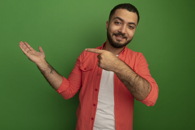 手指身穿橘色衬衫 留着胡须的年轻人 手上拿着食指指着一边 微笑着自信地站在绿色的墙上侧身目录胡须