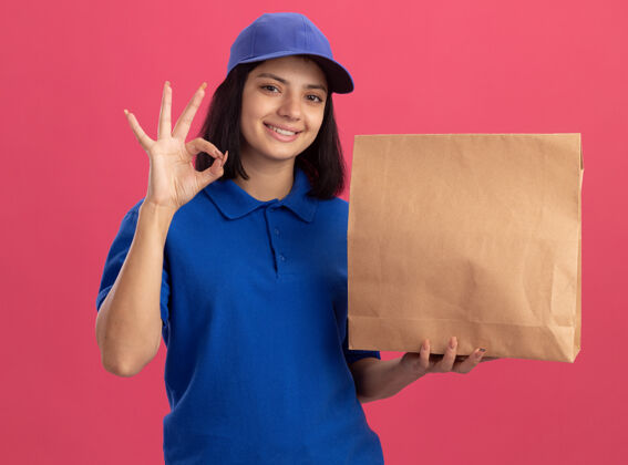 微笑身穿蓝色制服 头戴鸭舌帽 手持印有“ok”标志的纸包装的年轻送货员站在粉红色的墙上 开心地微笑着抱着女孩帽子