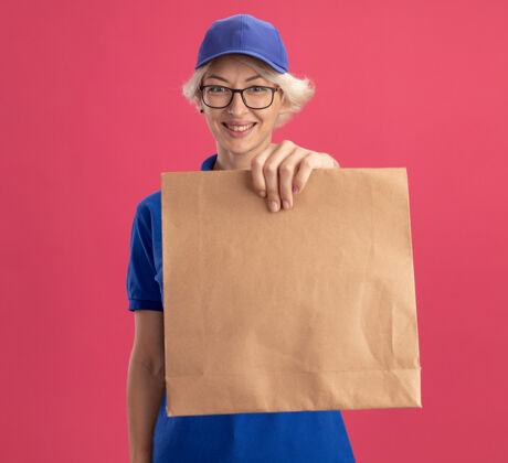 戴着身穿蓝色制服 戴着眼镜 拿着纸包的年轻送货员在粉红色的墙上欢快地微笑着欢呼帽子微笑