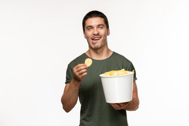 微笑正面图：穿着绿色t恤的年轻男性 带土豆cips和在白墙上吃饭的人孤独的电影成人电影土豆