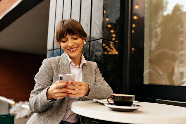 学习漂亮的快乐女士穿着时髦的衣服 用智能手机深褐色皮肤 穿着灰色夹克的女人坐在外面 拿着电话 咖啡和笔记本电脑集中女士青少年