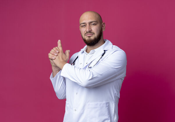 秃头年轻的秃头男医生穿着医用长袍 手持听诊器 在粉色背景上显示的姿势长袍医疗相机