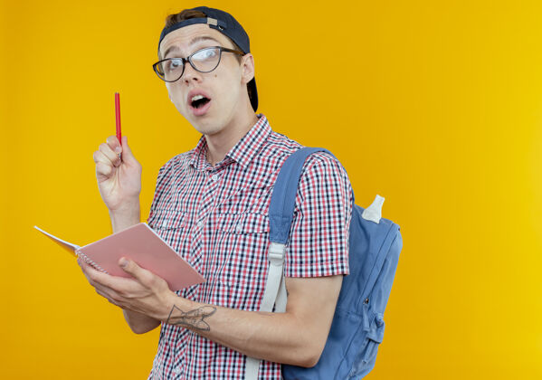 印象令人印象深刻的年轻学生男孩戴着背袋和眼镜和帽子拿着笔记本和钢笔隔离在白色背景上戴着拿着笔记本