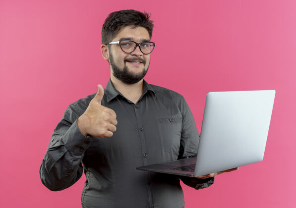 拇指戴着眼镜的年轻商人高兴地把拇指举在粉红色背景上年轻人眼镜笔记本电脑