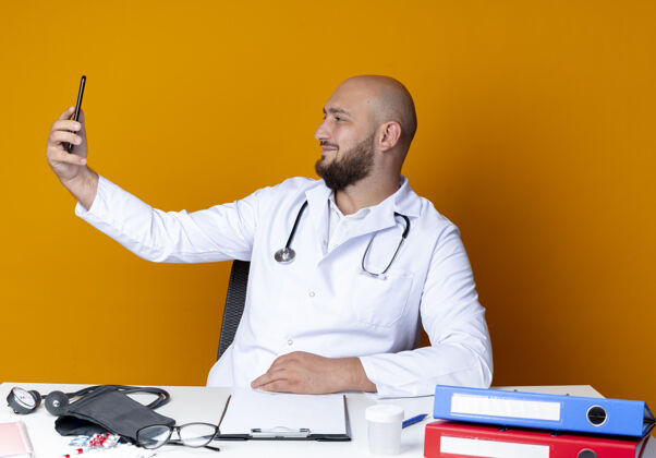办公桌高兴的年轻秃头男医生穿着医用长袍和听诊器坐在工作台上与医疗工具采取自拍隔离在橙色背景医疗自拍男性
