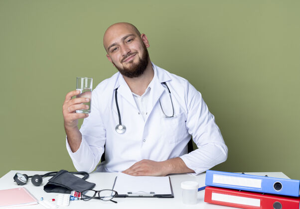 工作高兴的年轻秃头男医生穿着医用长袍和听诊器坐在办公桌前 拿着医疗工具拿着一杯隔离在绿色背景上的水抱着男性坐着