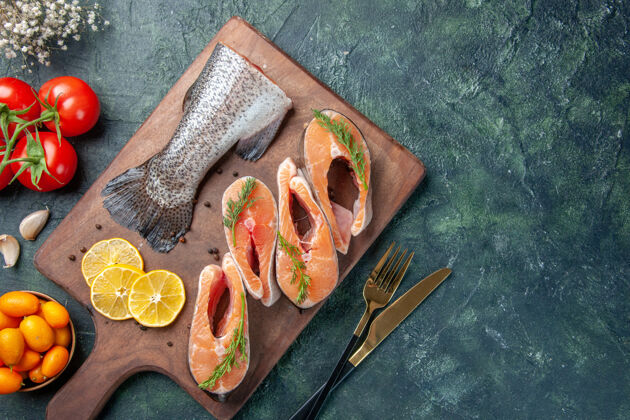 工具生鱼俯视图柠檬片绿色胡椒木砧板西红柿餐具放在黑暗的桌子上刀片刀食物