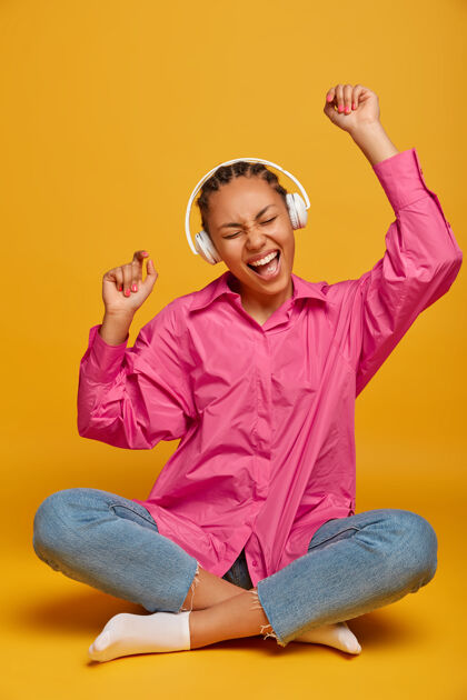 音乐开朗的年轻女子戴着耳机听音轨 举起双臂 以莲花的姿势靠着黄墙坐着 随着音乐的节奏移动 充满活力 感到快乐和放松人 休闲年轻音频民族