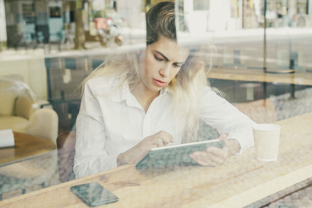 项目专注的女性专业人士坐在办公桌旁的共同工作空间或咖啡厅 使用平板电脑透过玻璃肖像沉思