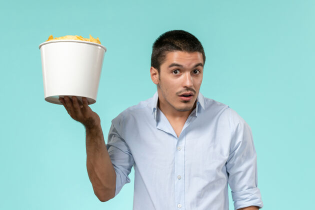 浅蓝色正面图年轻人拿着篮子和土豆cips在淡蓝色的墙上遥远的电影院孤独的男人微笑篮子容器