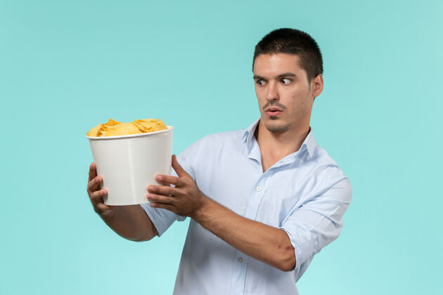 微笑前视图年轻人拿着篮子 在浅蓝色的墙上放着电影遥控器餐具杯子马克杯