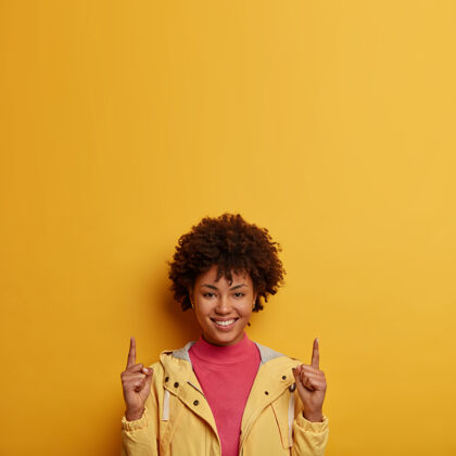 随意快乐微笑的黑皮肤女人用食指指着上面的垂直镜头 给出建议或建议 建议顺着这个方向 穿着夹克 隔离在黄色的墙上请积极高兴