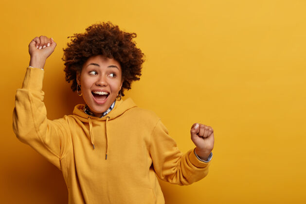 感动一张非洲裔美国女孩跳幸运舞的照片 在万岁中举手 获得胜利后感觉像冠军 快乐地注视着某处 玩得开心 感觉音乐的节奏 孤立在黄色的墙上满意姿势非洲