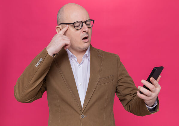 男人一个穿着西装戴眼镜的中年秃头男人站在粉红色的墙上看着他的手机 感到困惑和焦虑戴着电话站着