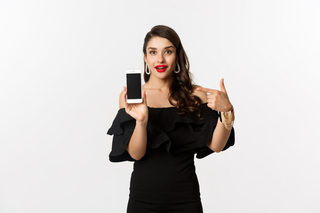 年轻网上购物概念穿着黑色连衣裙的时尚女性用手指着智能手机屏幕 展示应用程序 站在白色背景上服装优雅美丽