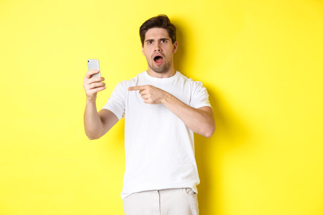 男性困惑的男人用手指着手机屏幕 看不懂什么 站在黄色背景上复制空间成年人移动年轻