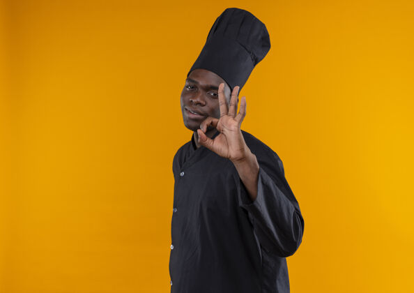 标志年轻自信的非洲裔美国厨师身着厨师制服 站在一旁 用手在橙色上做手势 并留有复印空间年轻手制服