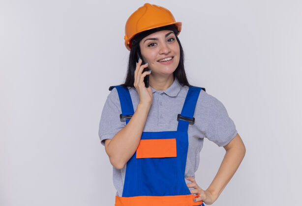 微笑身穿施工制服 头戴安全帽的年轻建筑工人站在白色的墙上 一边微笑着一边用手机交谈年轻人建筑工人手机
