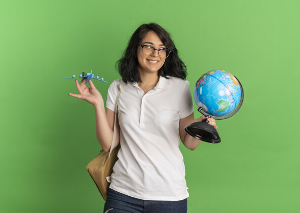 空间年轻的高加索女孩 戴着眼镜 背着书包 微笑着 拿着玩具飞机和地球仪 放在绿色的空间里包地球年轻