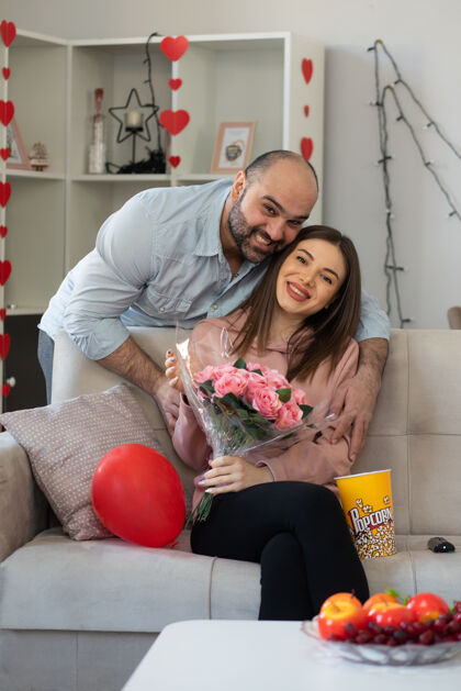 坐着年轻漂亮的情侣快乐的男人送一束鲜花给他微笑的女朋友坐在沙发上在灯光客厅庆祝国际妇女节微笑国际年轻