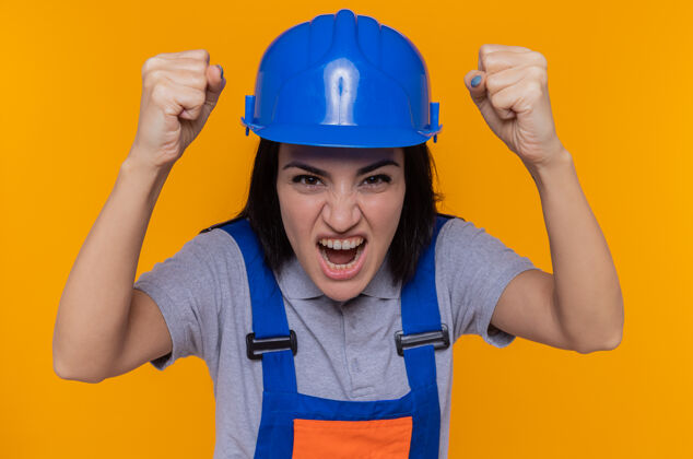 年轻愤怒的年轻建筑妇女身穿建筑制服 戴着安全帽 举起拳头 站在橙色的墙上 表情咄咄逼人地大喊大叫站立女人头盔