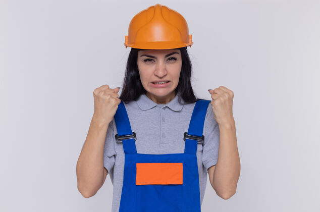 女人身穿建筑制服 头戴安全帽的年轻建筑工人站在白墙上 紧握拳头 愤怒而沮丧地看着前方愤怒制服安全