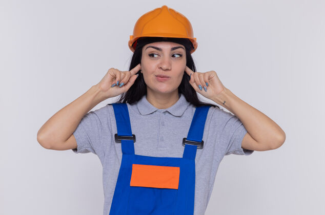 盖身穿建筑制服 戴着安全帽 用手指捂住耳朵的年轻建筑工人站在白色的墙壁上 看上去很困惑安全女人制服