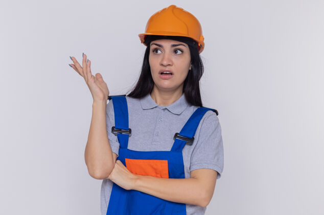 不高兴身穿建筑制服 头戴安全帽的年轻建筑妇女站在白色的墙上 困惑地 不高兴地把胳膊伸到一边看手臂年轻建设者