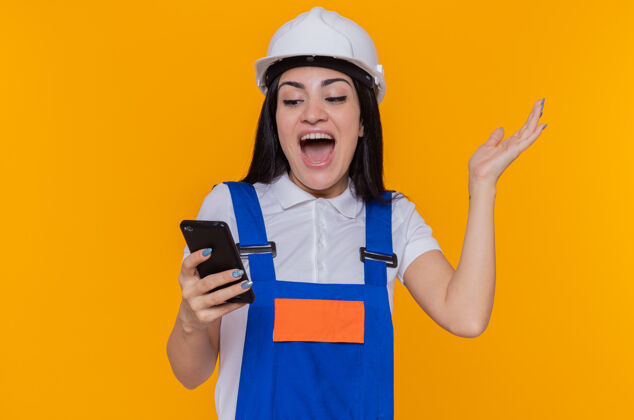 建筑身穿建筑制服 戴着安全帽 手持智能手机的年轻建筑工人站在橘色的墙上 兴奋而快乐地看着它头盔快乐女人