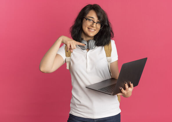 点年轻漂亮的白人女学生戴着耳机戴着眼镜 背着书包 指着粉色的笔记本电脑 还有复印空间穿脖子复制