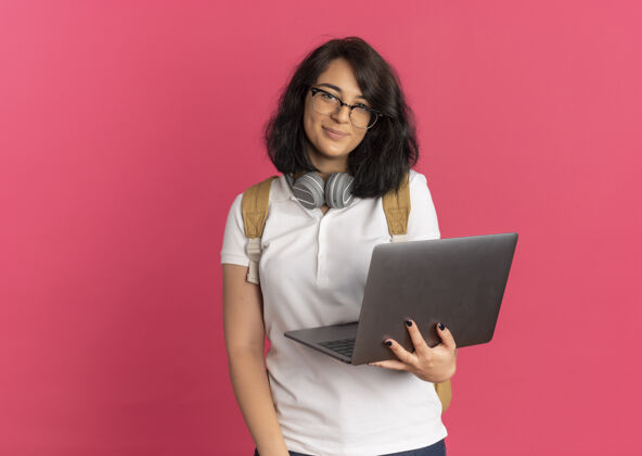 穿年轻可爱的白人女学生戴着耳机戴着眼镜背着包拿着笔记本电脑看着粉色的相机还有复印空间粉色耳机空间