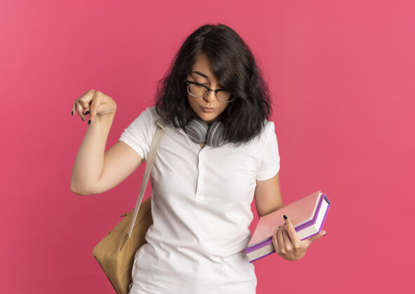 空间年轻漂亮的白人女生戴着眼镜 背着一个戴着耳机的袋子 看起来很惊讶 她拿着粉红色的书指着下面 留着复印空间女学生背部穿