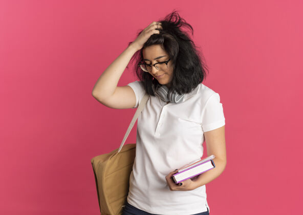 年轻年轻烦躁漂亮的白人女学生 戴着耳机 戴着眼镜 背着书包 低着头 拿着粉红色的书 还有复印空间心烦头复制