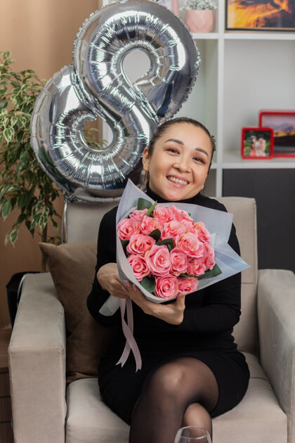 女人快乐的亚洲年轻女子身穿黑色连衣裙坐在椅子上 手里拿着一束鲜花 在灯光明媚的客厅里欢快地笑着庆祝国际妇女节生活花束花