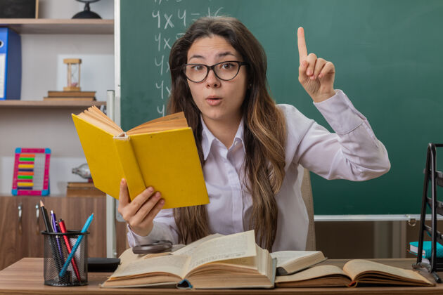 前面年轻的女老师戴着眼镜 带着书在教室里看书 惊讶地伸出食指坐在课桌前的黑板前女人学校坐着