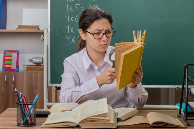 黑板戴眼镜的年轻女教师坐在教室黑板前的课桌旁 用放大镜看书 感到困惑和不快不高兴女人放大镜