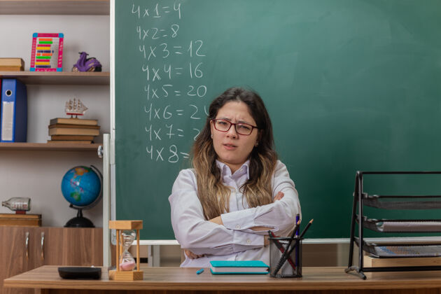 老师年轻的女老师戴着眼镜看着前面困惑和不高兴的双臂交叉着坐在课桌前的教室黑板前困惑书桌穿