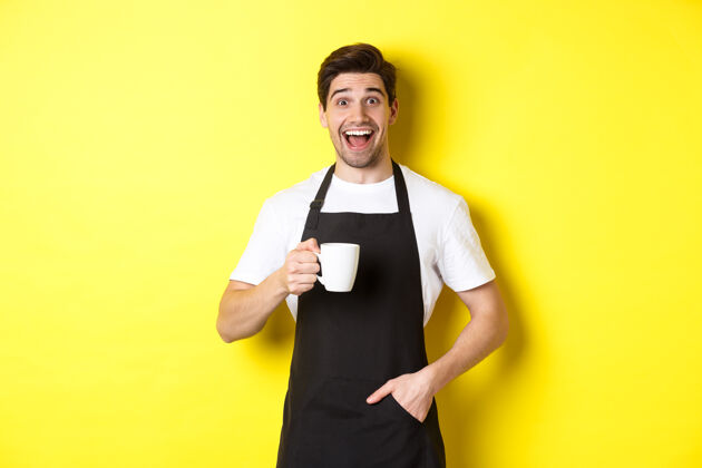 咖啡师微笑着的年轻咖啡师 穿着黑色围裙 手里拿着咖啡杯 站在黄色背景上围裙模特帅哥