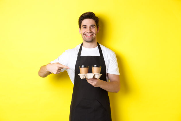 男士帅气的咖啡师拿着两杯外卖咖啡 指着饮料微笑着 站在黄色背景下的黑色围裙里咖啡黄色表情