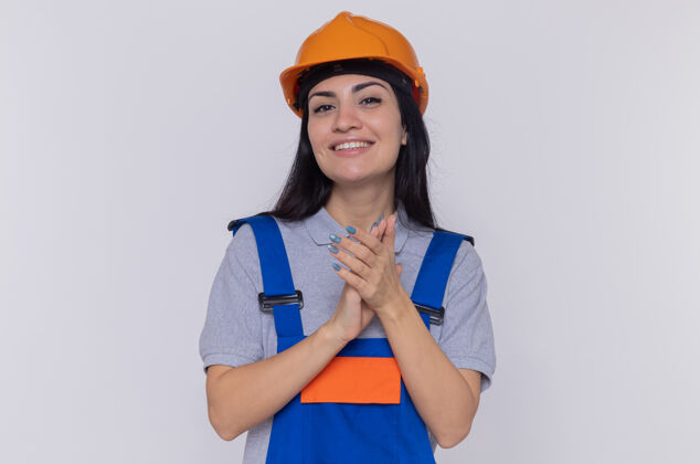 抱着身穿施工制服 头戴安全帽的年轻建筑工人手牵着手 面带微笑自信地站在白色的墙壁上看着前方女人建设者头盔