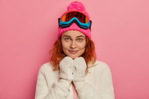套头衫迷人的红发女人戴着暖和的帽子 滑雪镜和白色套头衫女人衣服滑雪