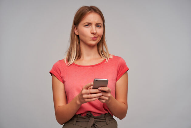 学生长相可疑的女人 金发的成年女孩 穿着粉色t恤和棕色裙子 手持智能手机不信任 隔着灰墙孤立着电话皮肤年轻