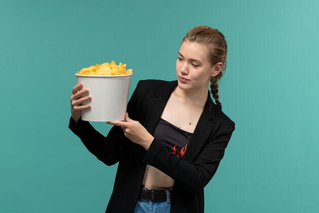 抱着正面图：年轻女性拿着薯片包 在浅蓝色的表面上看电影包装电影院年轻女性