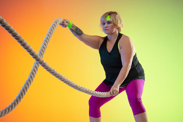 力量年轻的白人大号女模特在霓虹灯下的橙色渐变背景上训练用绳子做锻炼运动的概念 健康的生活方式 积极的身体 平等拉伸运动运动员