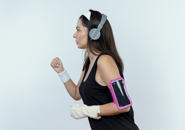 女士戴着头戴耳机和智能手机袖标的年轻健身女士站在白墙上锻炼站立健身工作