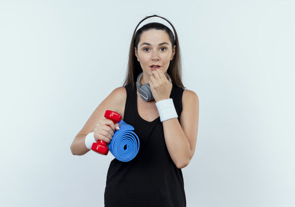 站着戴着头巾 手持哑铃和瑜伽垫的年轻健身女士惊讶地站在白墙上抱着健身耳机